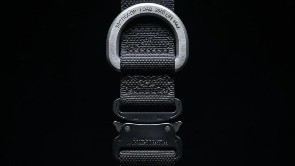 Close-up details of a pet harness featuring a rugged, utilitarian "tech-wear" aesthetic; set against a solid black background and illuminated by a cool-white spotlight, the image highlights a substantial matte-finish metal D-ring, a high-strength engineering-grade plastic buckle, and bold, rugged nylon stitching.